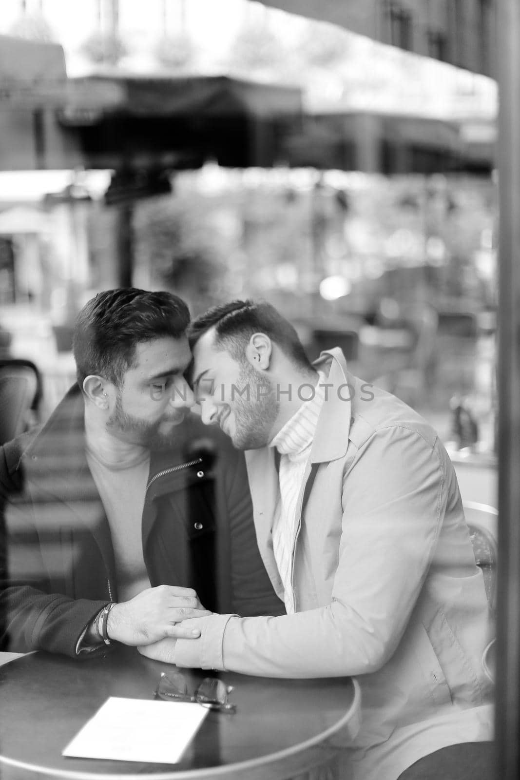 Black and white photo of caucasian gays sitting at cafe and hugging, reflection in glass. by sisterspro