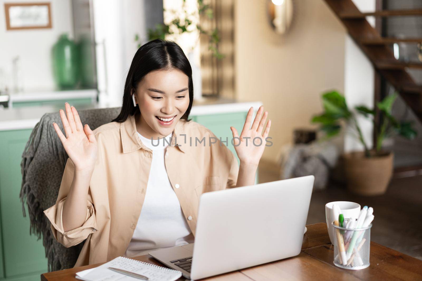 Asian girl on video conference, saying hello, waving hand at laptop camera, working remote from home. Young woman connects to webinar or online classes.
