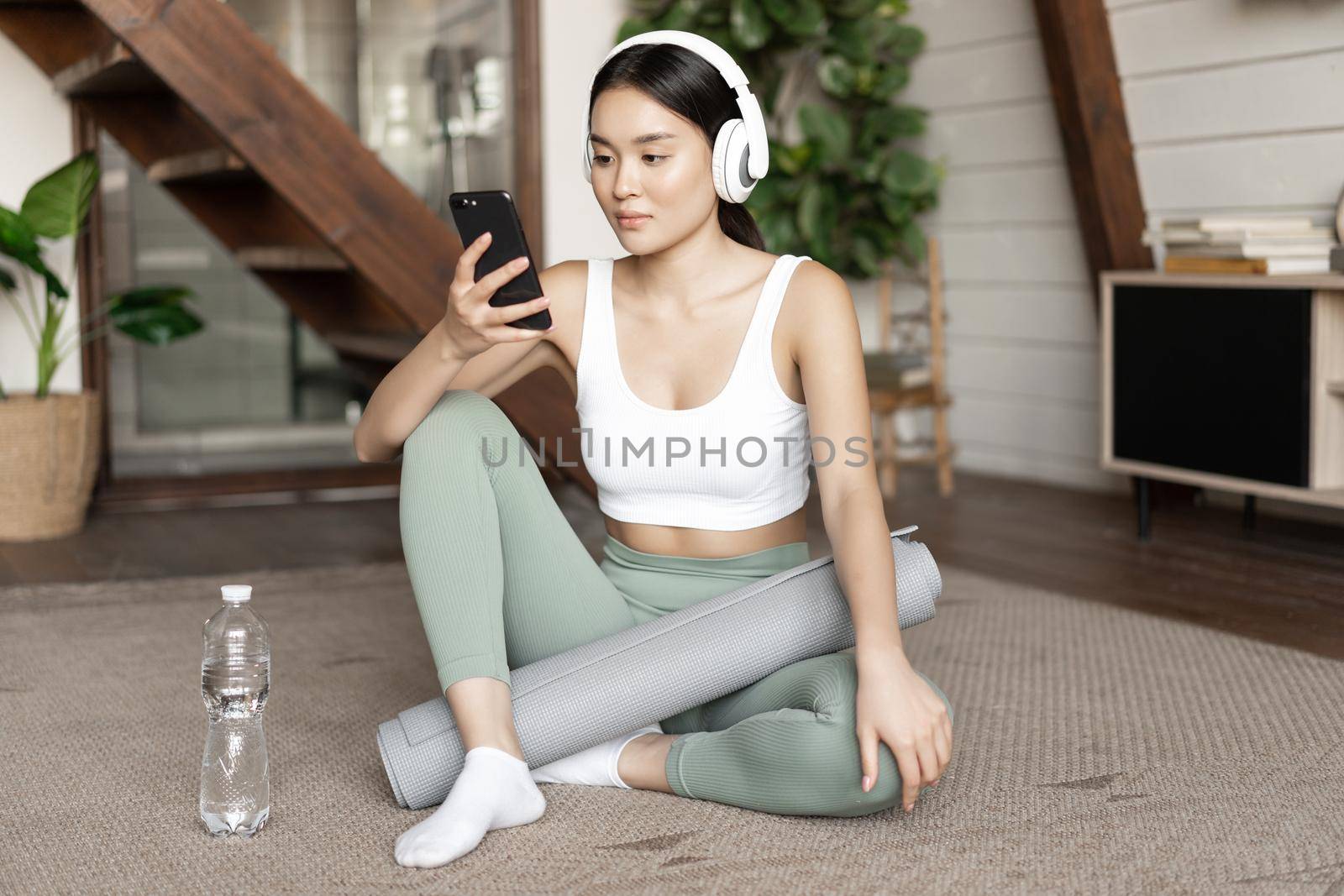 Young asian woman with water bottle, listening music or podcast in headphones during fitness at home, sitting on floor before exercising by Benzoix