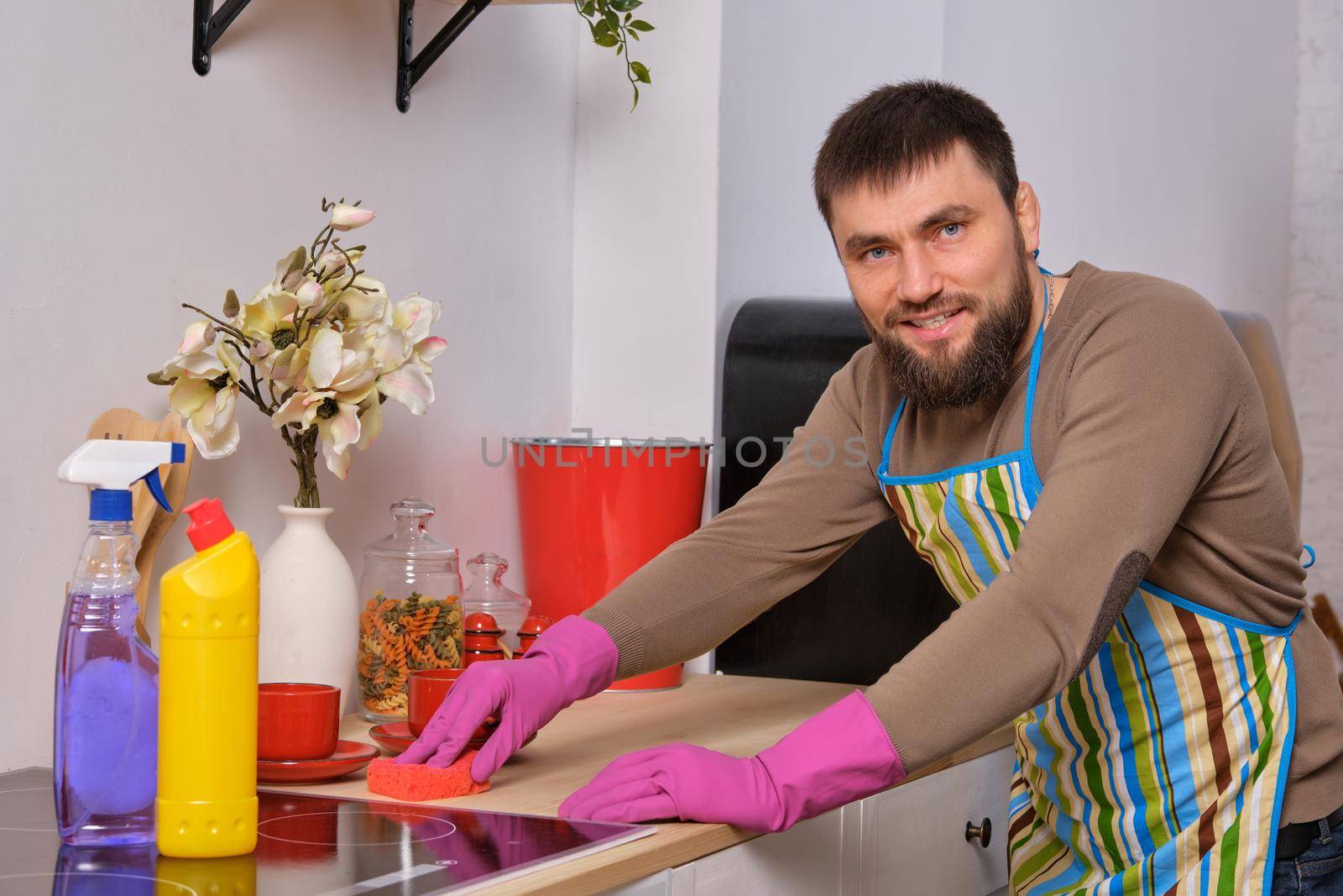 Image of a young Caucasian bearded man in the kitchen wearing apron. He tries to clean the cooking surface and near. Different detergents used
