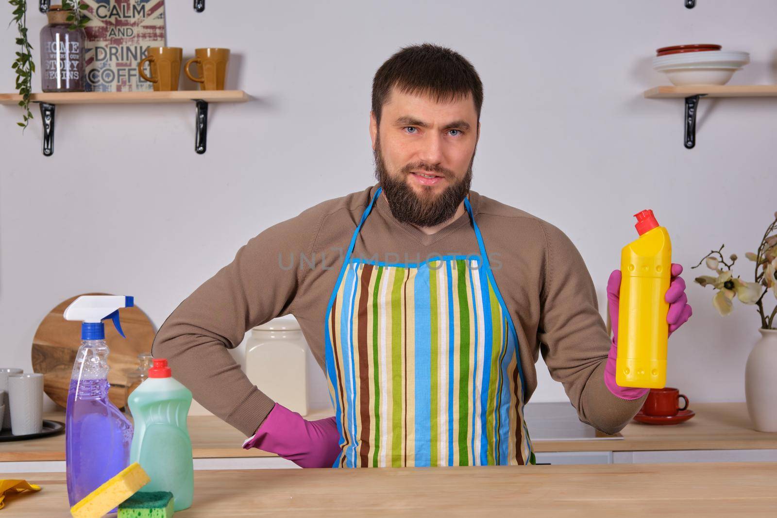 Housecleaning concept. Young bearded man in kitchen makes some advertising. He promotes detergent, holding it in one hand and shows at camera