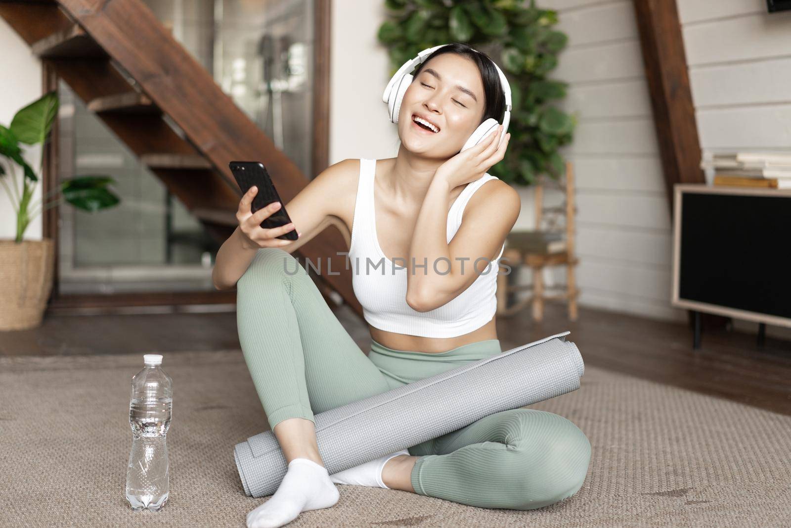 Happy asian girl in fitness clothing sitting at home on floor, listening music and workout, holding smartphone and singing during training session.