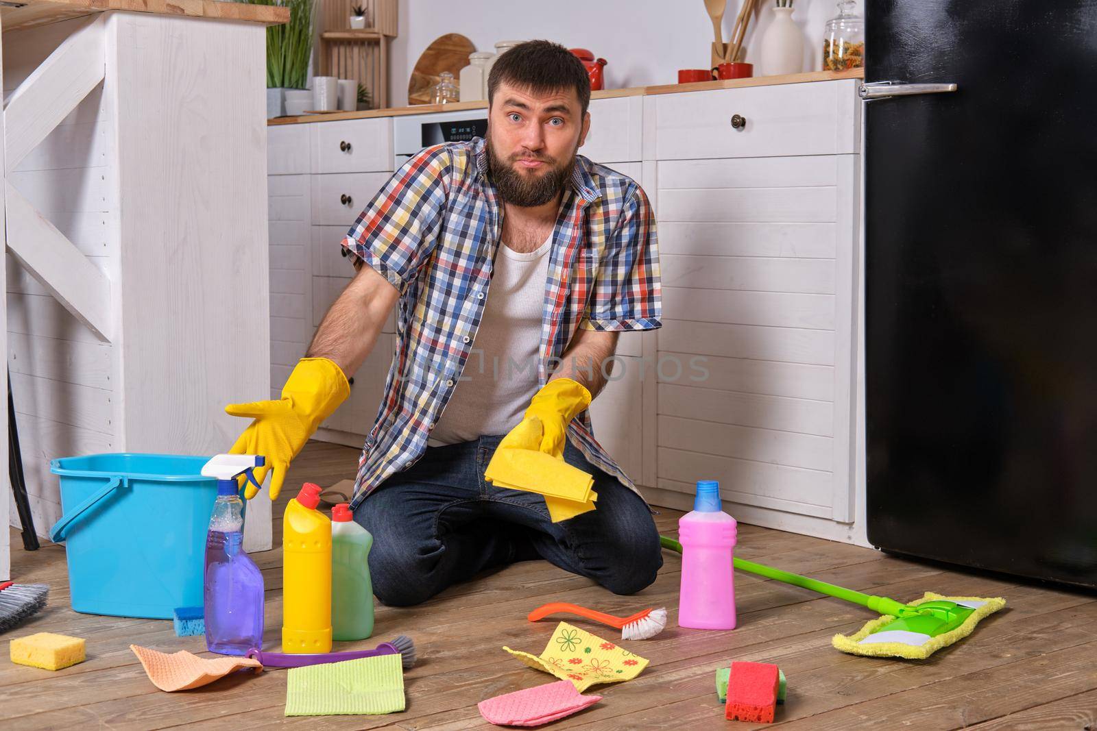 Housecleaning concept. Caucasian handsome young bearded man in checkered shirt sits on the floor of his kitchen and tries to clean it using all of his detergents, rags, washcloths and brushes