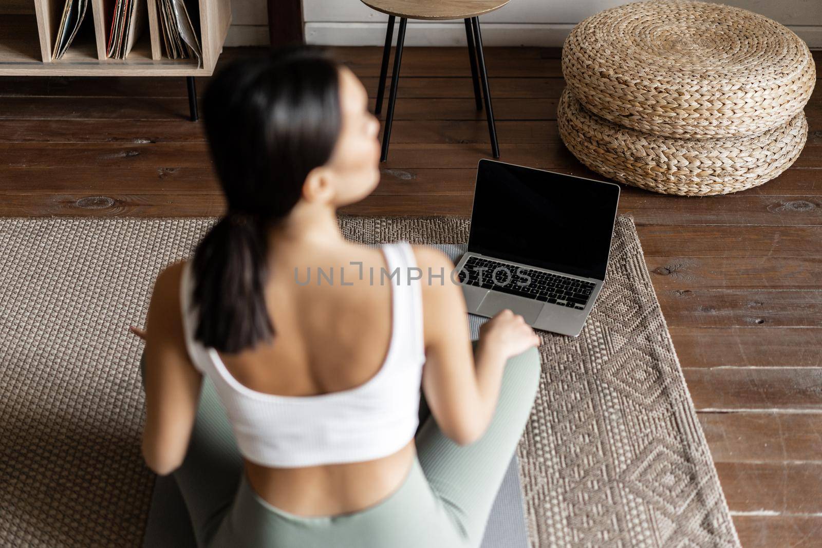 Mindfulness and meditation concept. Rear view of young asian woman meditating at home, following online course on laptop, sitting in living room on floor mat by Benzoix