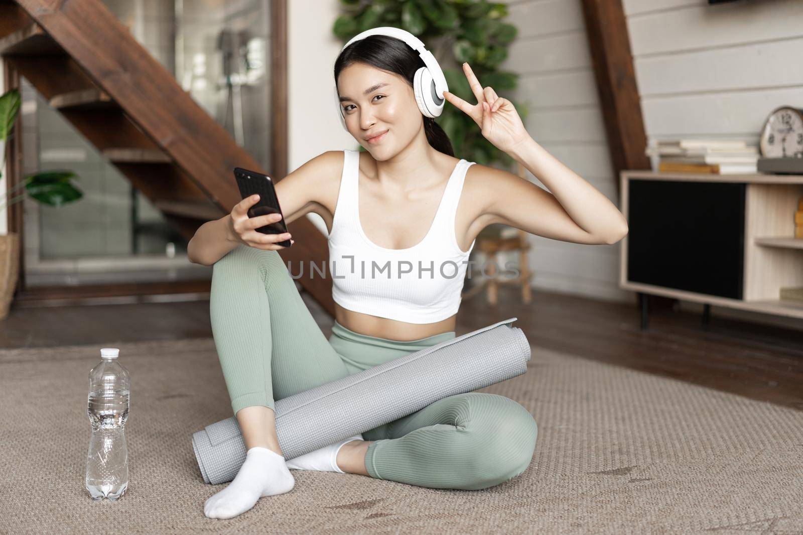 Happy sporty girl sitting on floor at home and taking selfie during workout, wearing headphones and holding smartphone. Concept of sport and lifestyle