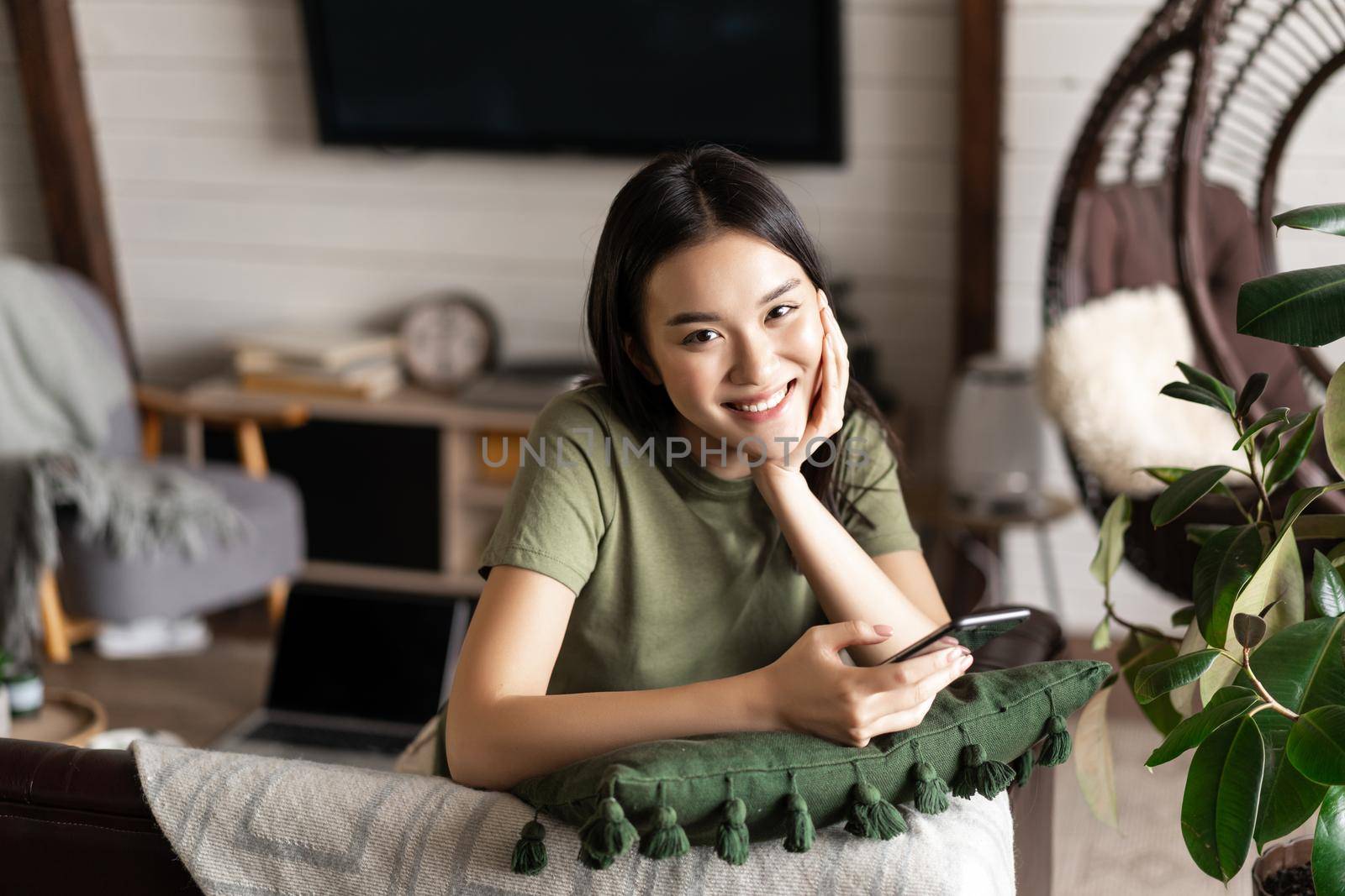 Smiling korean girl enjoying her weekend at home, resting at home on sofa, using mobile phone application, waiting for meal delivery.