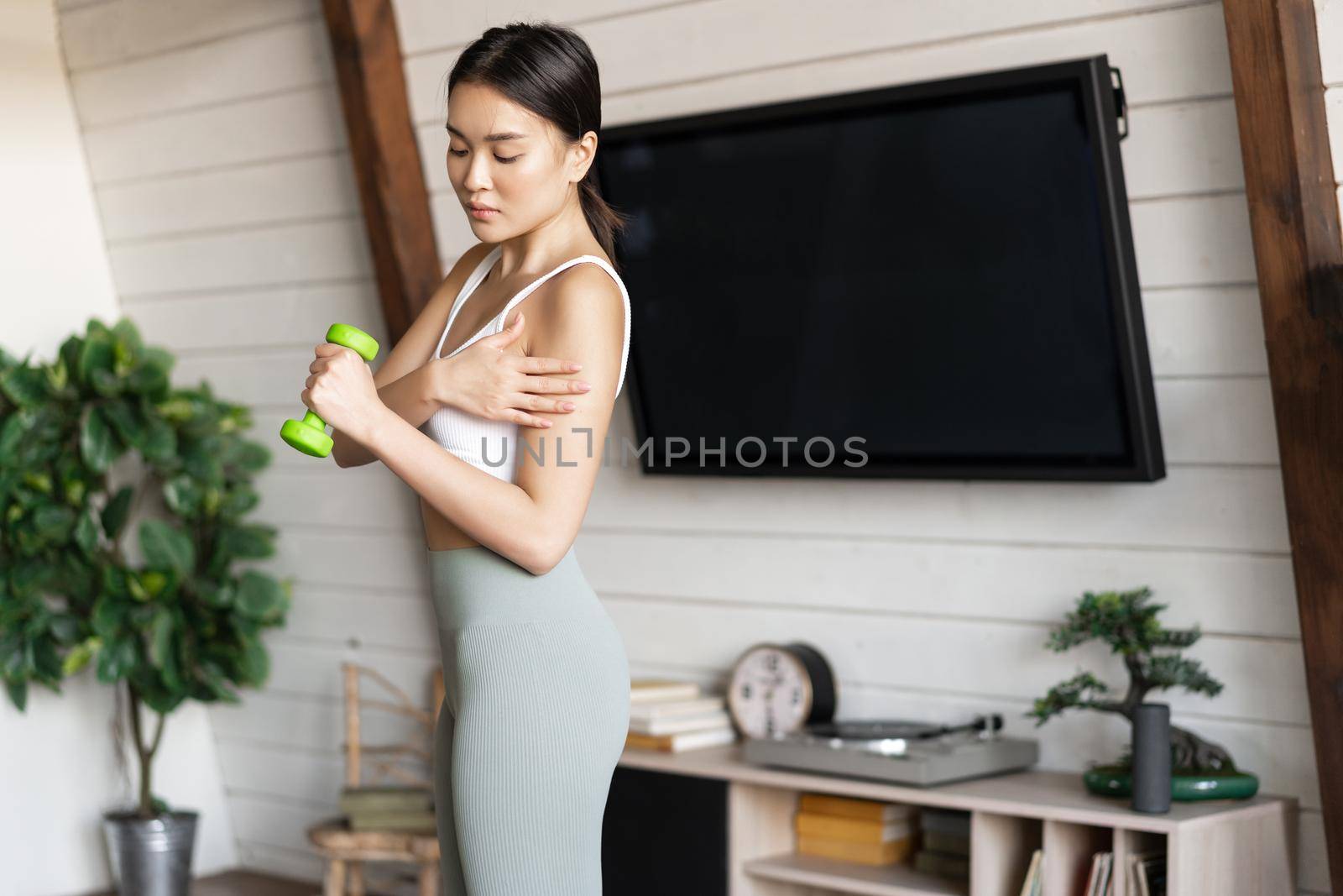 Active and healthy asian girl with fit body doing fitness exercises at home, lifting dumbbells and touching her biceps, workout in living room.