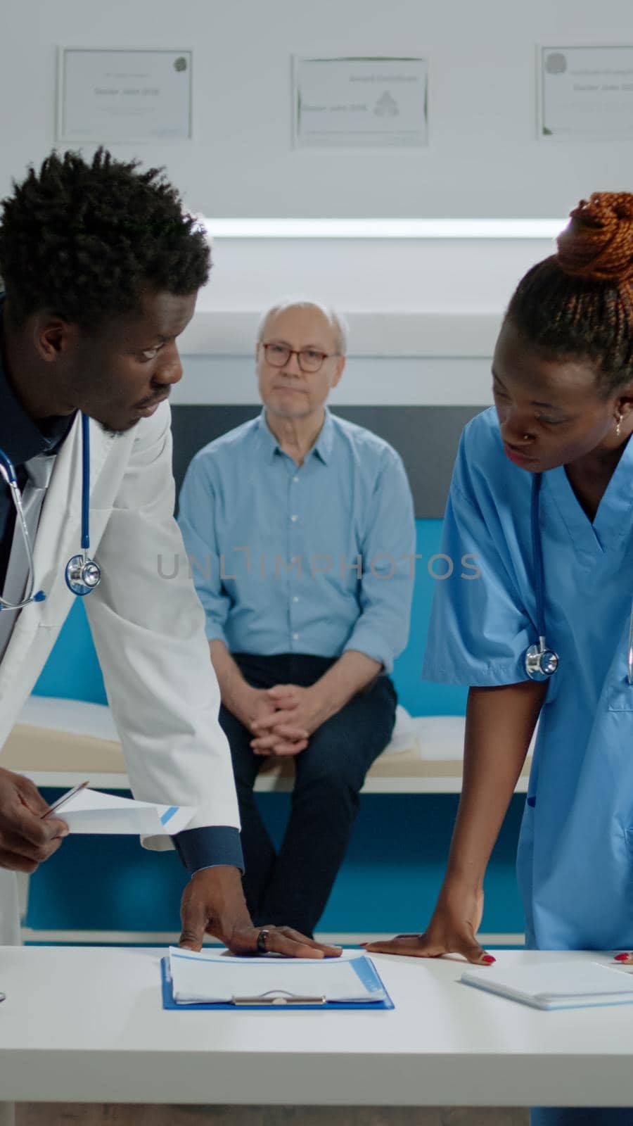 Medical team of people using instruments for checkup appointment with senior patient sitting on bed in background. Doctor and nurse with laptop and document files on desk for diagnosis