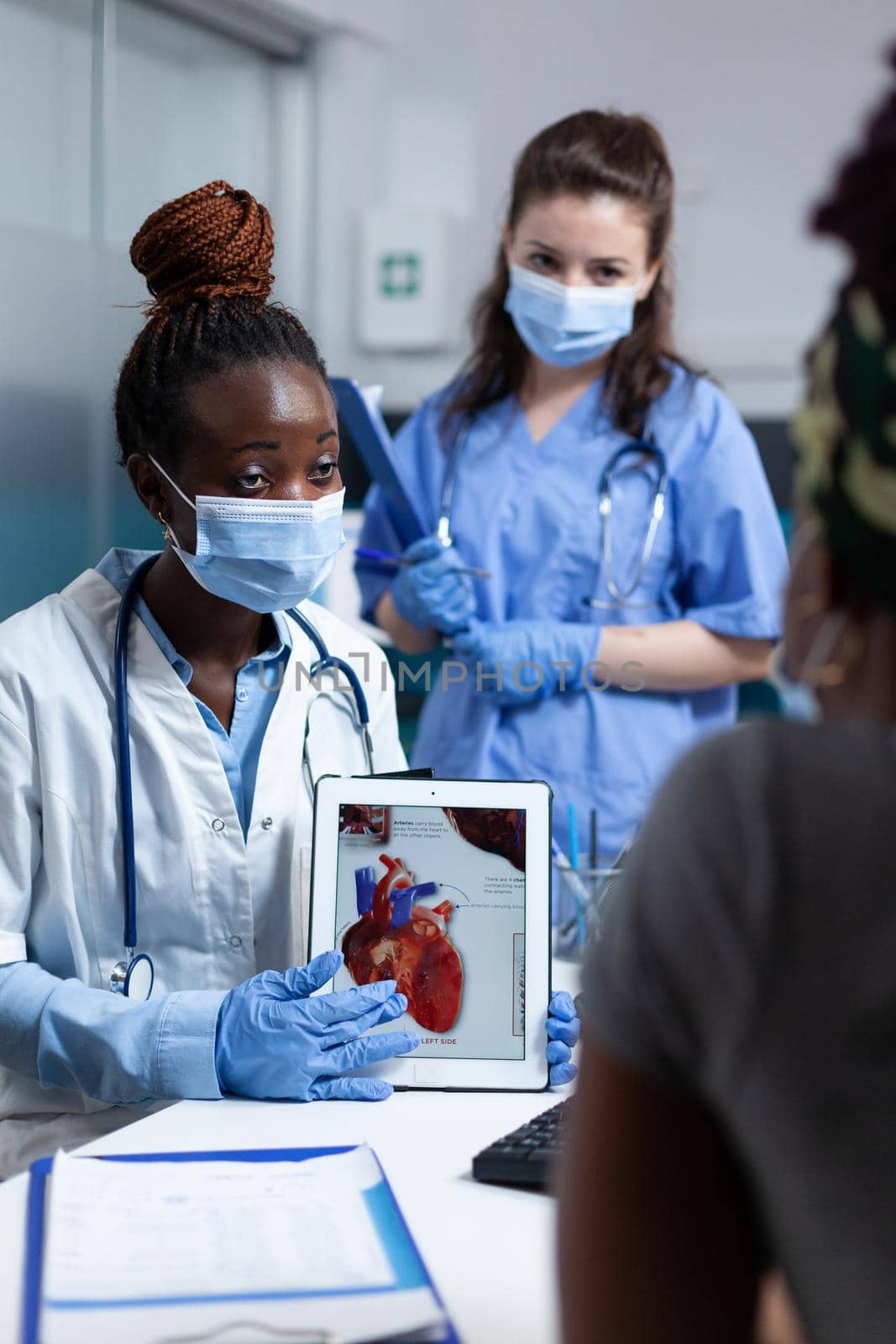 African american cardiologist doctor holding tablet showing heart radiography to sick patient explaining medicaton treatment during clinical appointment in hospital office. People with medical face mask