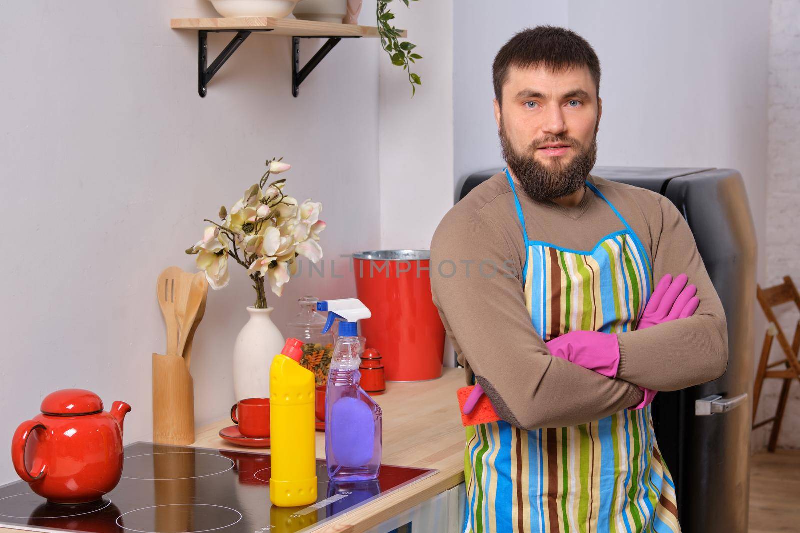 Image of a young Caucasian bearded man in the kitchen wearing apron. He tries to clean the cooking surface and near. Different detergents used