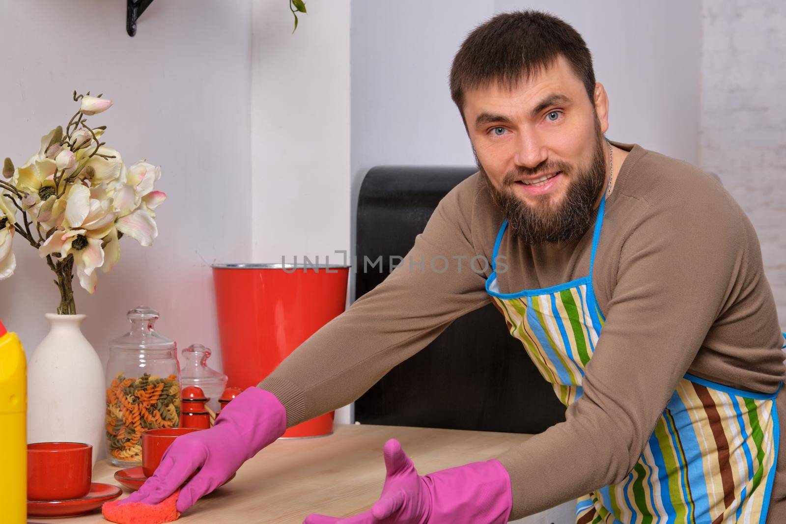 Image of a young Caucasian bearded man in the kitchen, he is desperately trying to clean a table, using detergent
