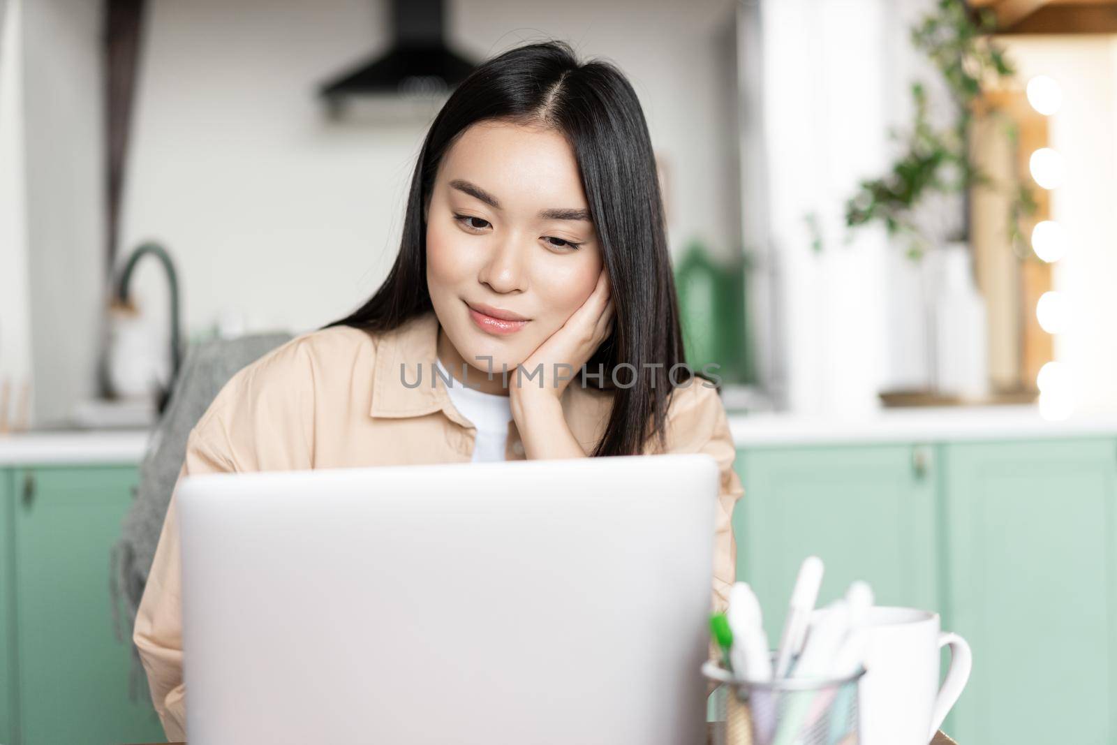 Smiling asian woman relaxing watching webinar, looking at laptop while sitting at home by Benzoix