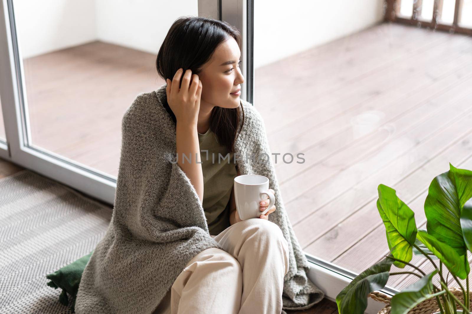 Young romantic asian woman enjoying weekend, relaxing morning with cup of tea, sitting on floor near window and looking outside. Lifestyle concept by Benzoix