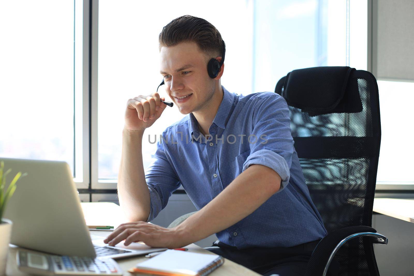 Cheerful young support phone male operator in headset, at workplace while using laptop, help service and client consulting call center concept.