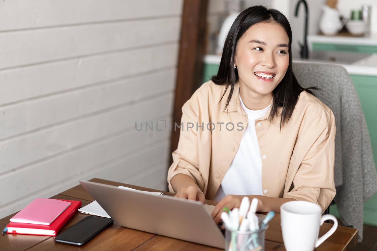 Young asian woman working from home on laptop, studying or doing homework with computer pc, sitting at kitchen table and smiling aside by Benzoix