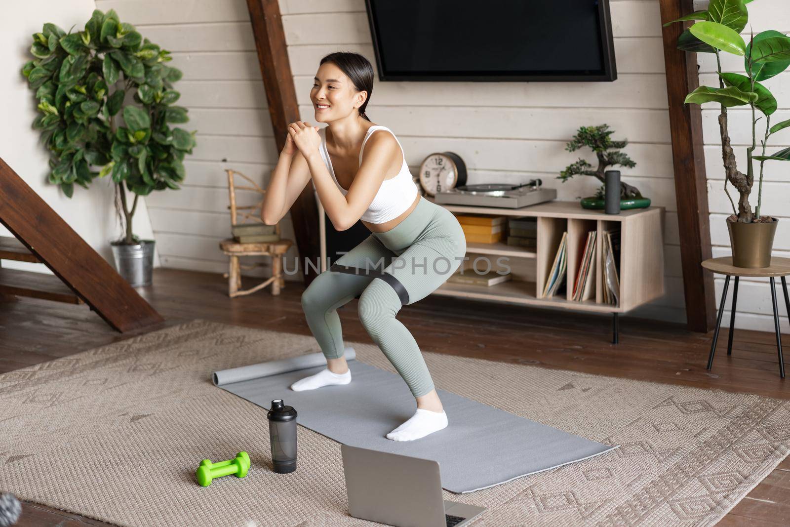 Cute asian fitness girl at home doing workout, squats with stretching elastic rope on legs, standing on floor mat, listening to online sport instructor on laptop video.