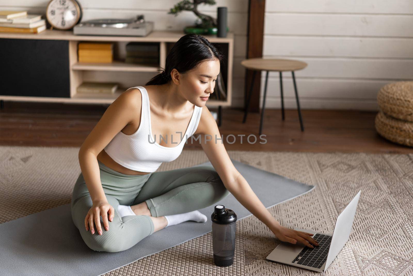Sport and lifestyle concept. Smiling young asian woman doing yoga at home, follow meditation course on laptop, sitting in zen pose on floor mat.