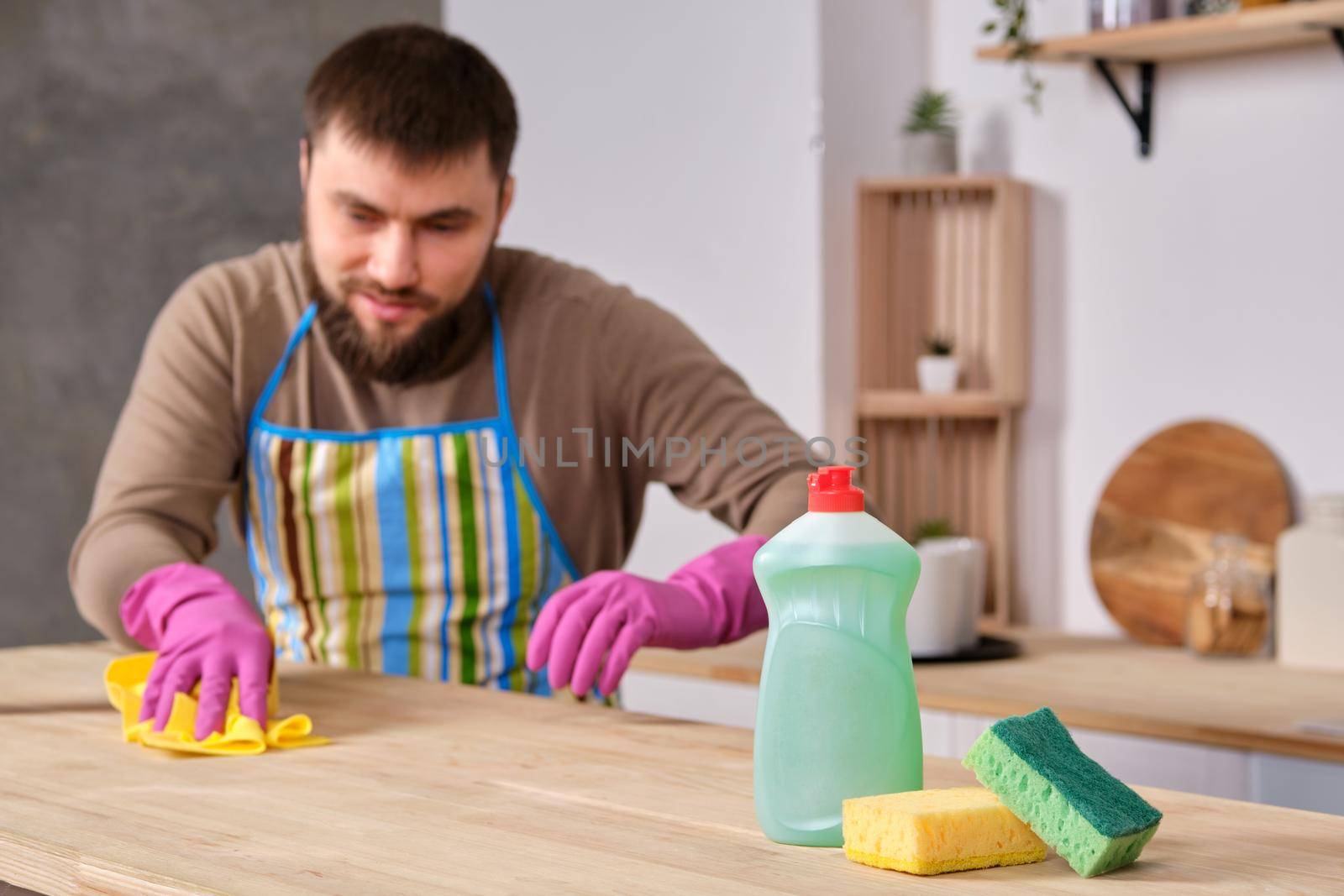 Image of a young Caucasian bearded man in the kitchen, he is desperately trying to clean a table, using detergent