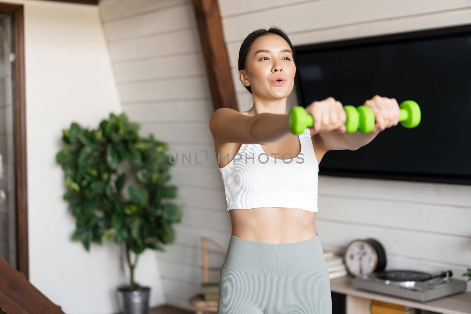 Fitness asian girl doing workout at home in living room, lifting dumbbells, wearing activewear for sport activities.