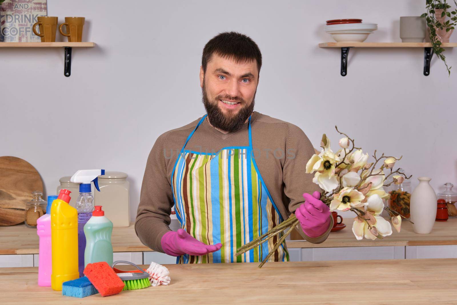 Image of a young Caucasian bearded man in the kitchen, he makes little presentation of all his detergents, preparing for big cleaning. Man caring for flowers