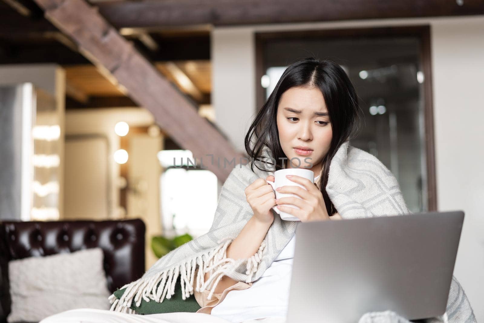 Image of sad asian girl looking at her laptop, being sick or catching cold, holding cup of tea. Concept of remote education, pandemic and quarantine.
