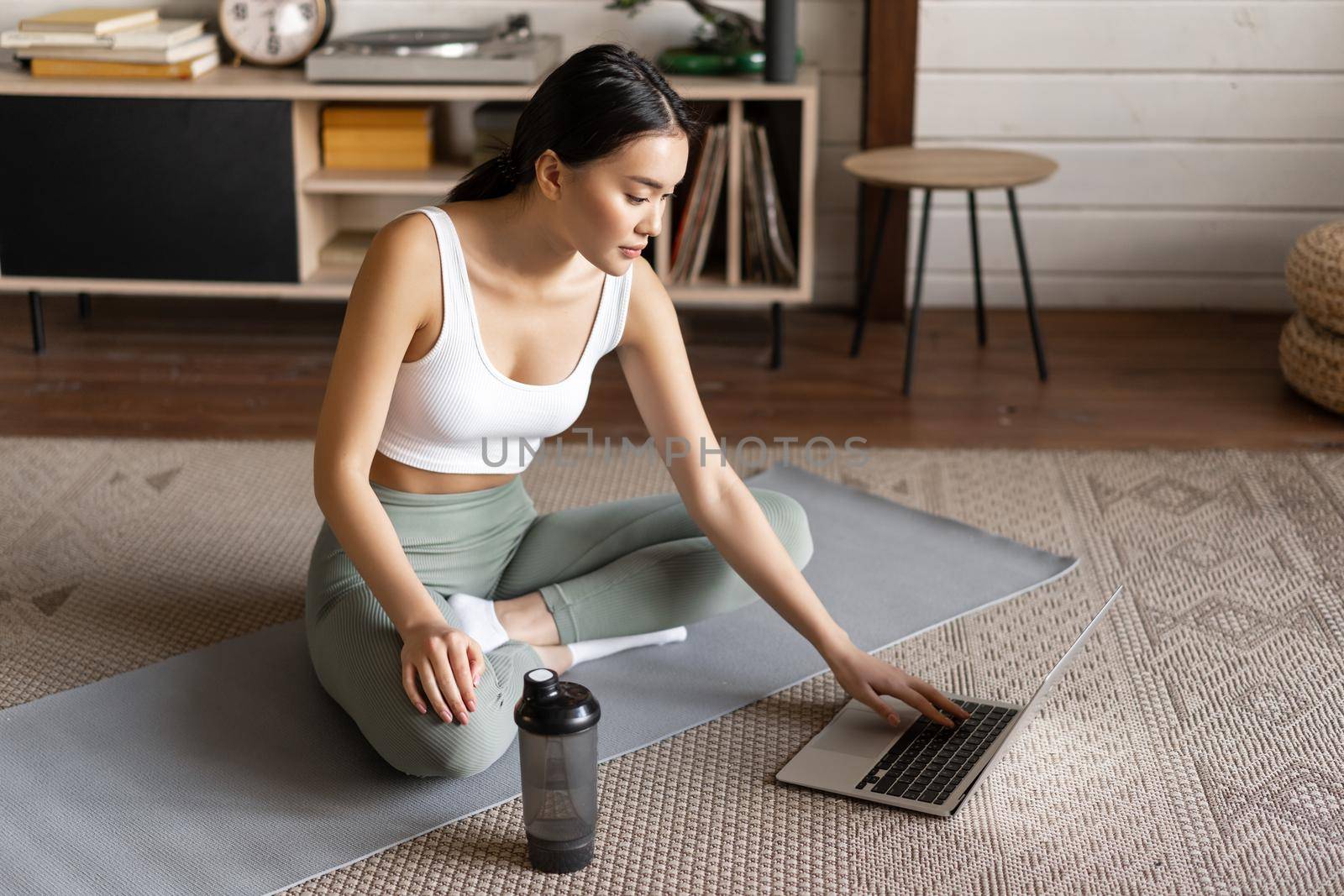 Sport and lifestyle concept. Smiling young asian woman doing yoga at home, follow meditation course on laptop, sitting in zen pose on floor mat.
