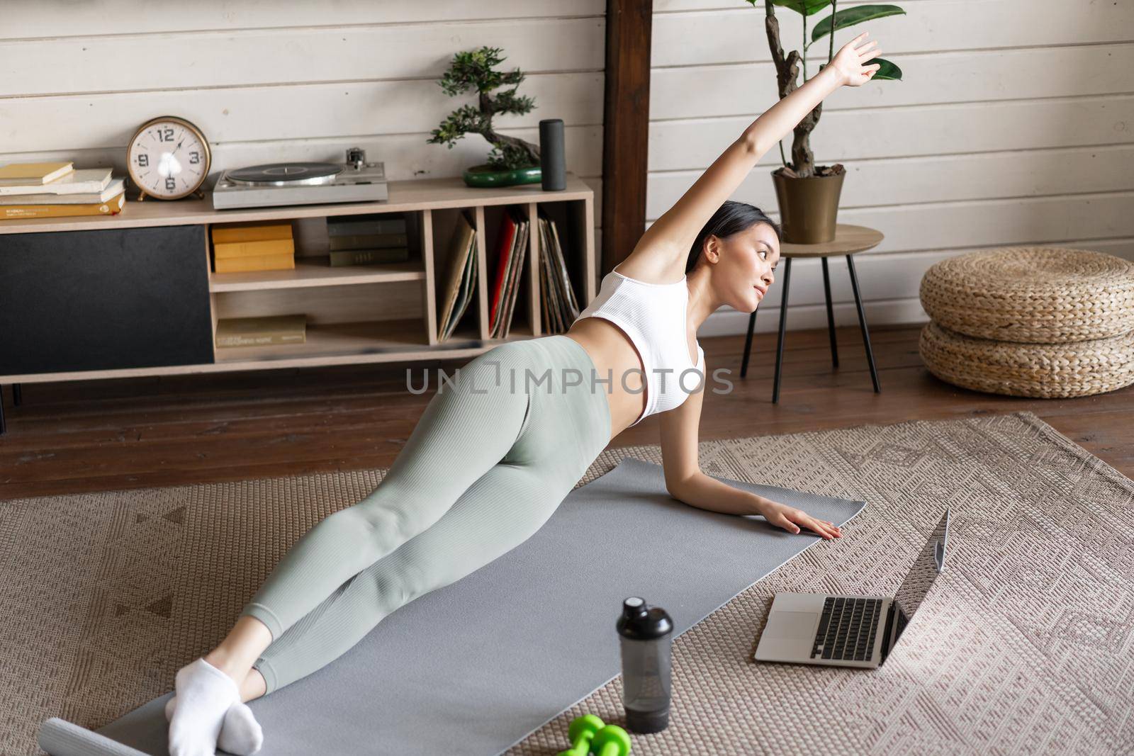 Young asian woman doing workout at home on floor mat, following sport instructor on laptop video, stretching arm and doing plank by Benzoix