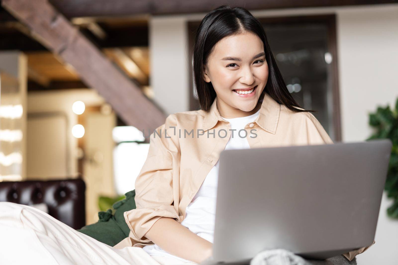 Smiling asian girl using laptop computer at home, freelancer working remote in living room, laughing at camera.