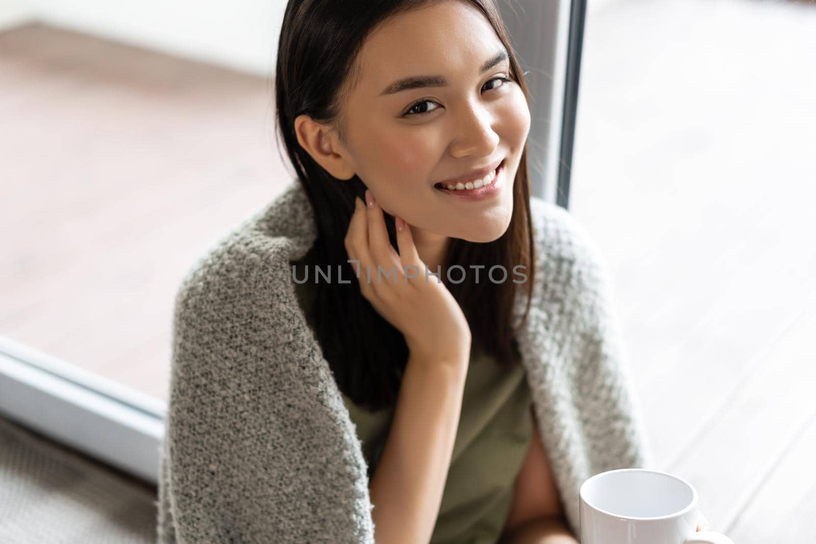 Portrait of beautiful asian woman wrapped in blanket, drinking coffee and smiling, relaxing at home on weekend, sitting near window and enjoying the view by Benzoix