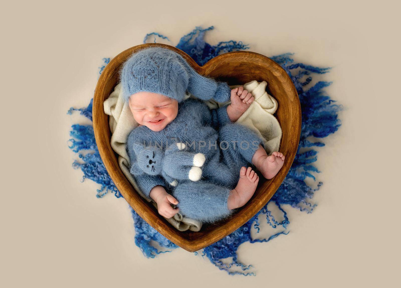 Newborn baby boy smiling in heart shape bed with knitted cat toy. Adorable infant child kid emotions during studio photoshoot wearing hat and costume