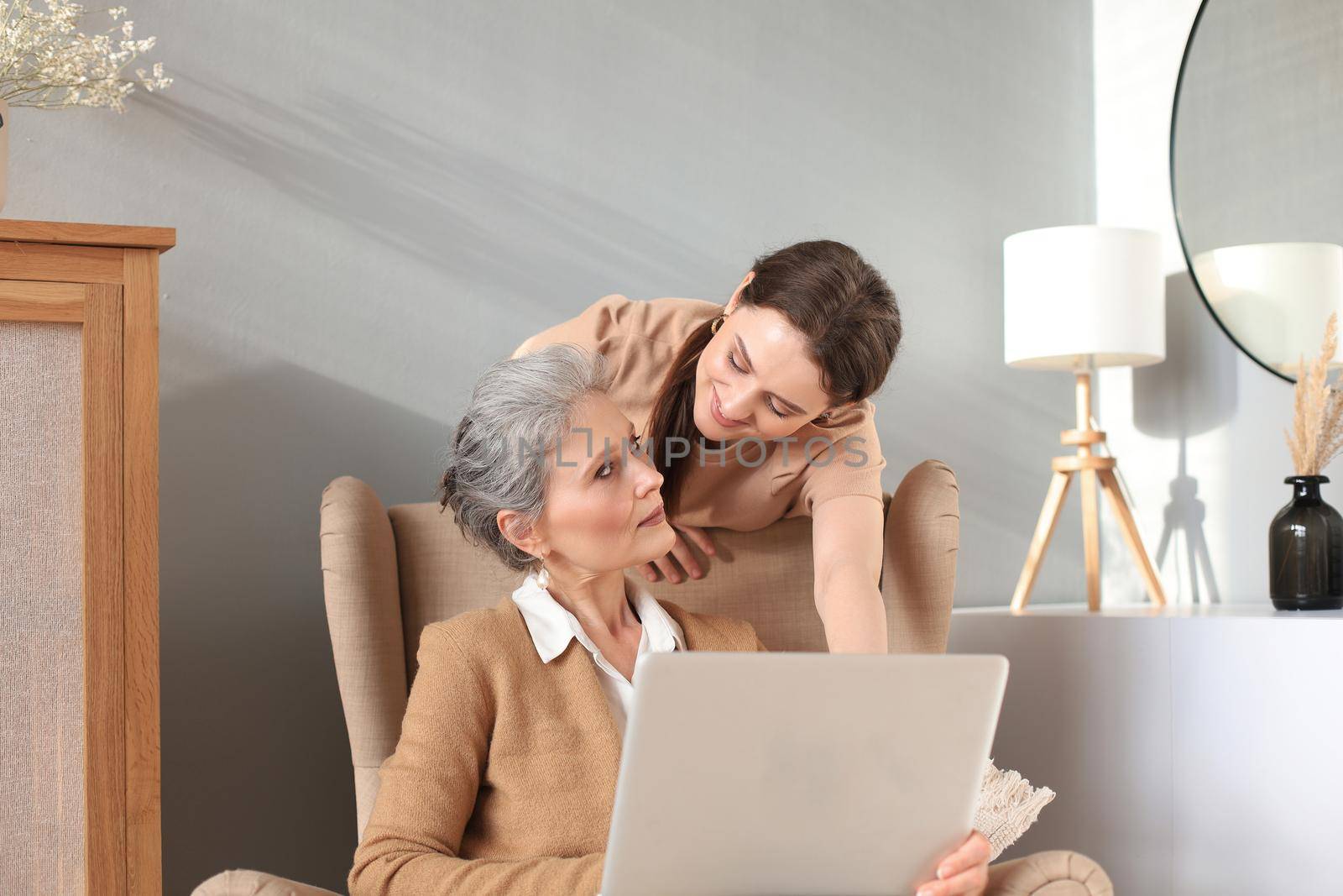 Happy elderly middle mother sitting on chair with her daughter, looking at laptop. Young woman showing video, photos to mommy, trusted relations. Family concept