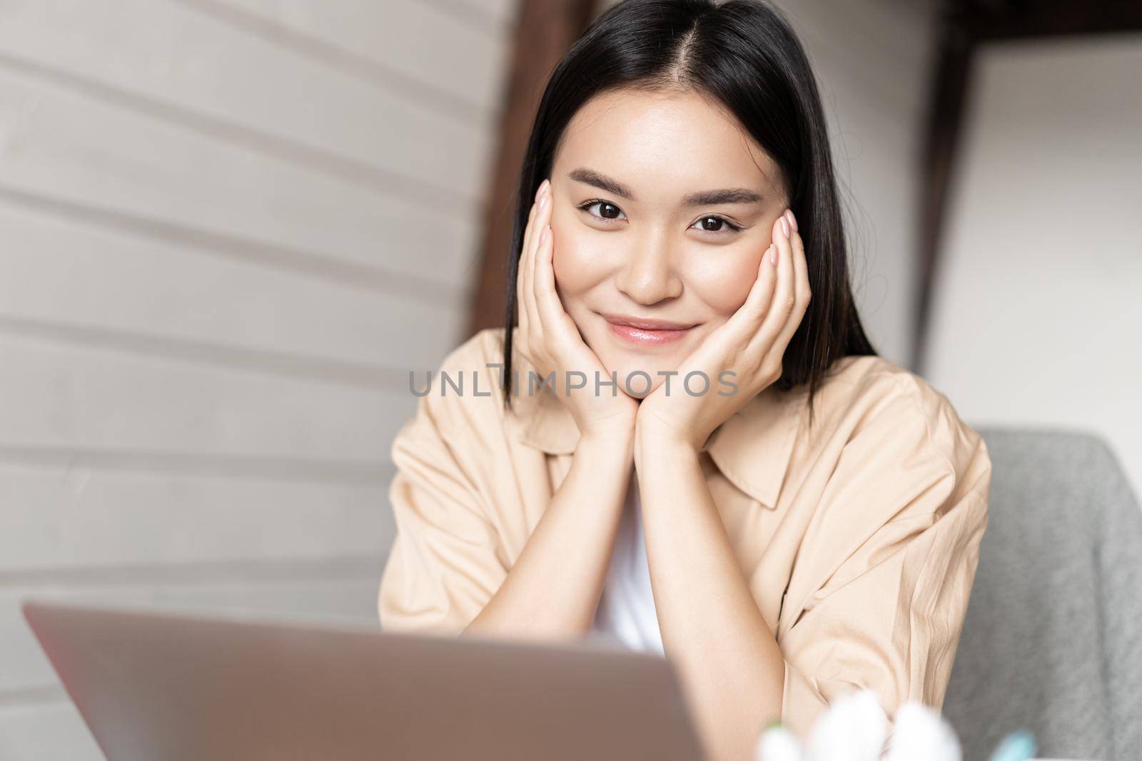 Portrait of asian woman smiling, working on laptop from home, resting with computer, sitting relaxed in kitchen near pc.