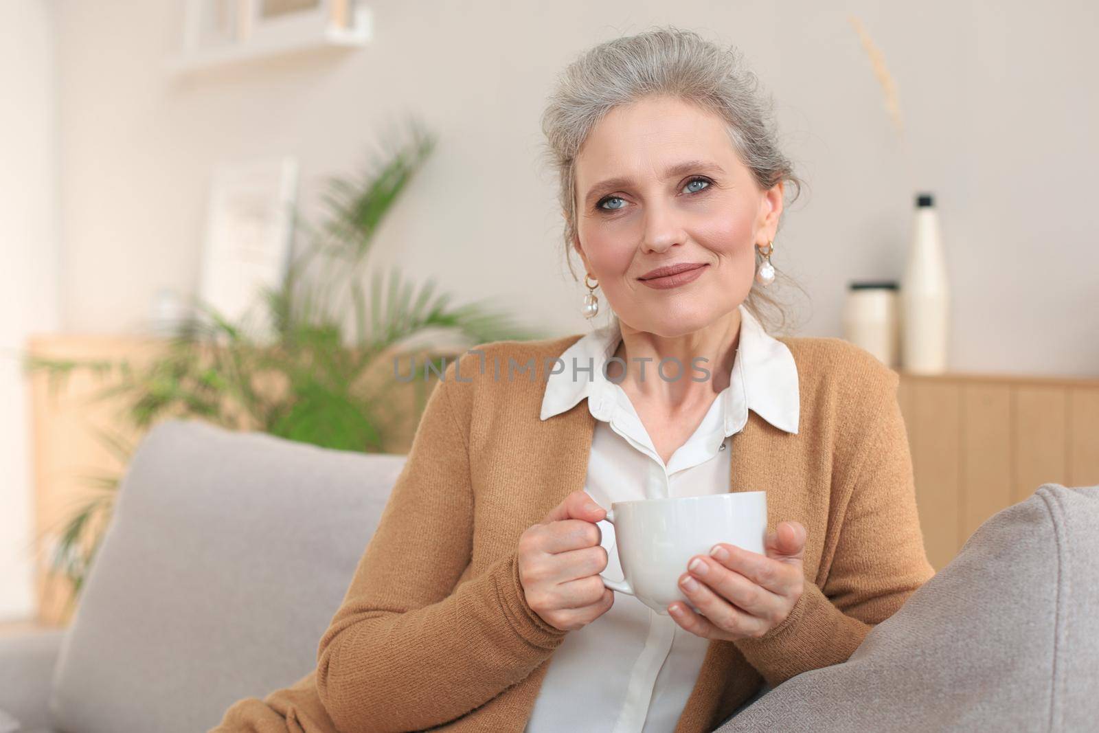 Happy mature woman resting on comfortable sofa drink coffee or tea, looking away, relaxing on cozy couch at home enjoy hot beverage