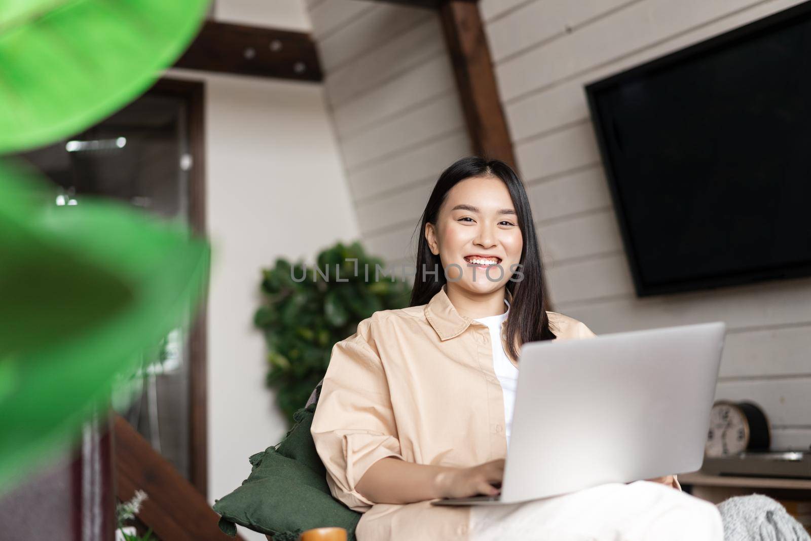 Smiling korean girl sitting and resting at home near tv, using laptop, relaxing and surfing net.