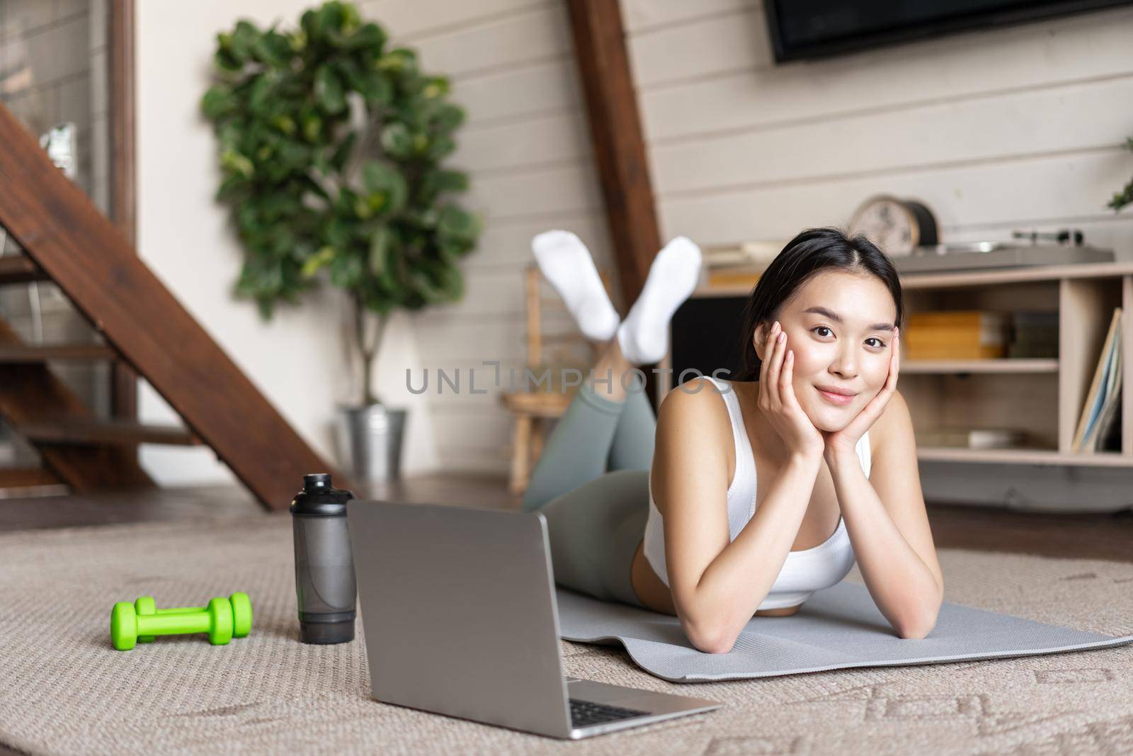 Asian fitness girl taking online fitness course, watching sports video, exercising at home on floor mat with dumbells and protein shaker bottle, smiling at camera.