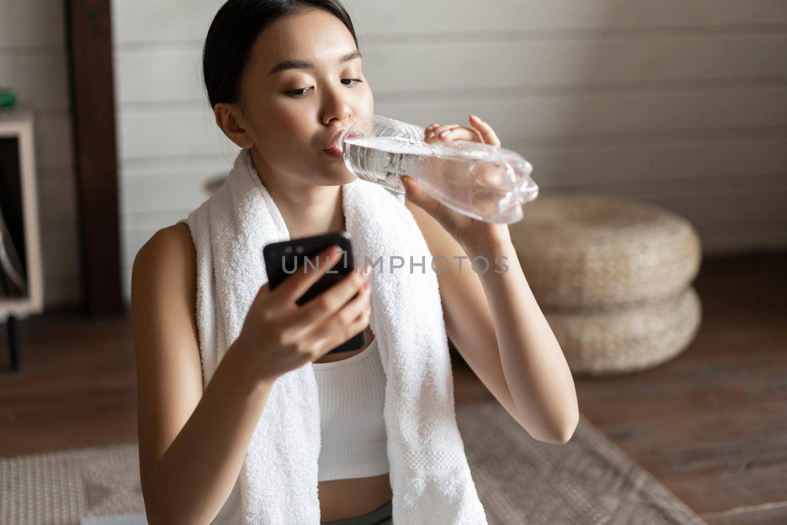 Image of young asian fitness girl with fit body, towel wrapped around neck, looking at mobile phone app, checking application during workout by Benzoix