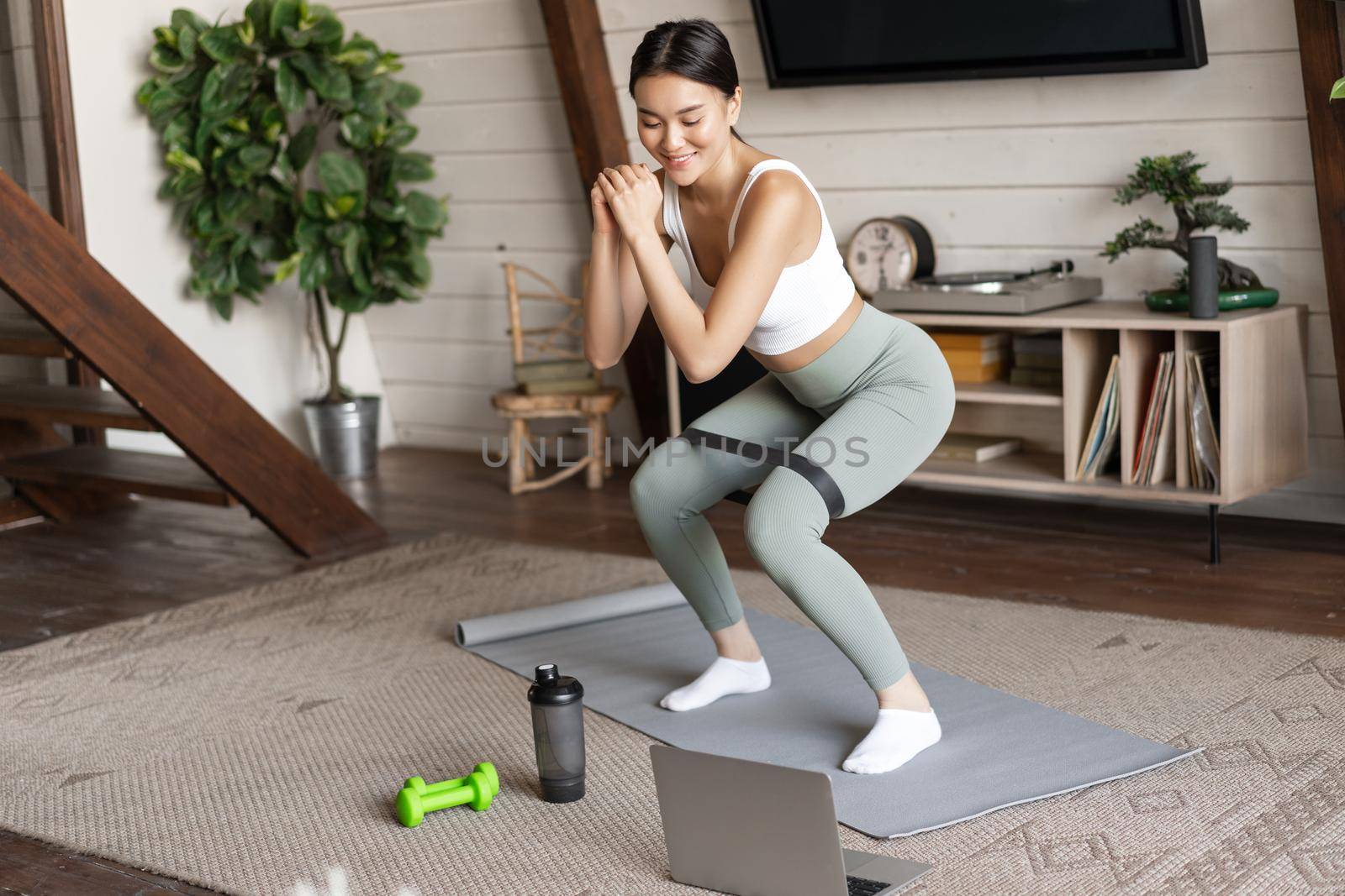 Cute asian fitness girl at home doing workout, squats with stretching elastic rope on legs, standing on floor mat, listening to online sport instructor on laptop video by Benzoix