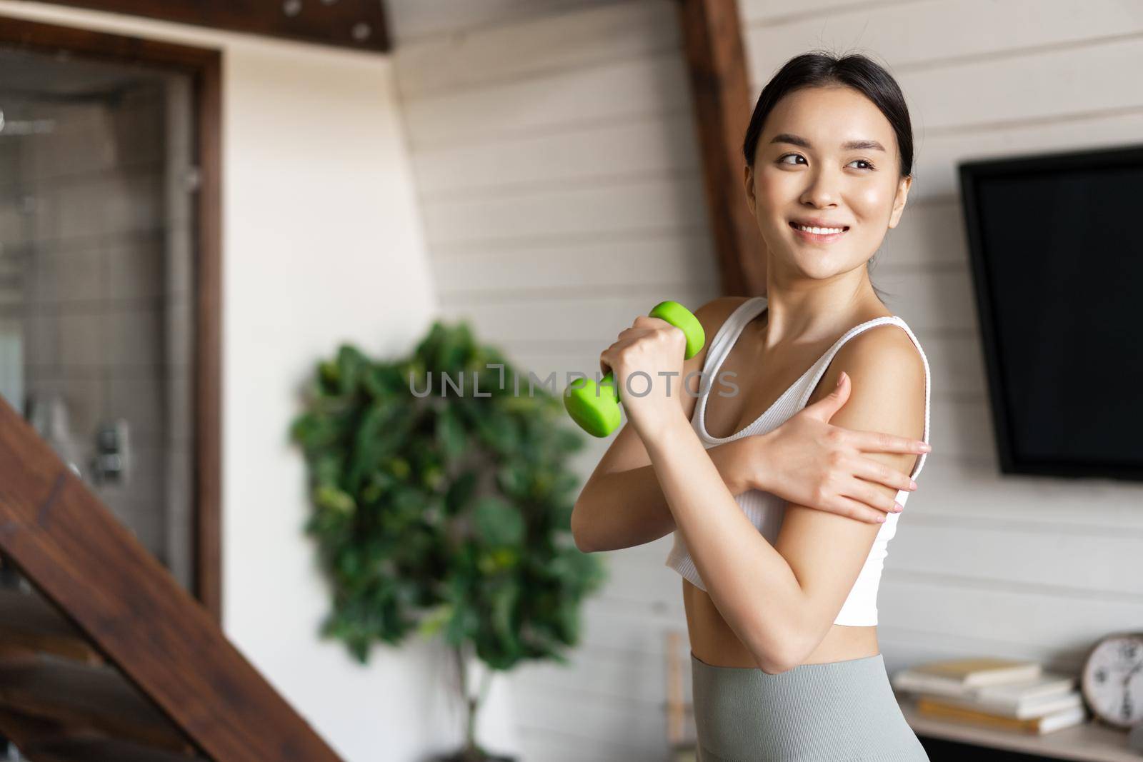 Active and healthy asian girl with fit body doing fitness exercises at home, lifting dumbbells and touching her biceps, workout in living room by Benzoix