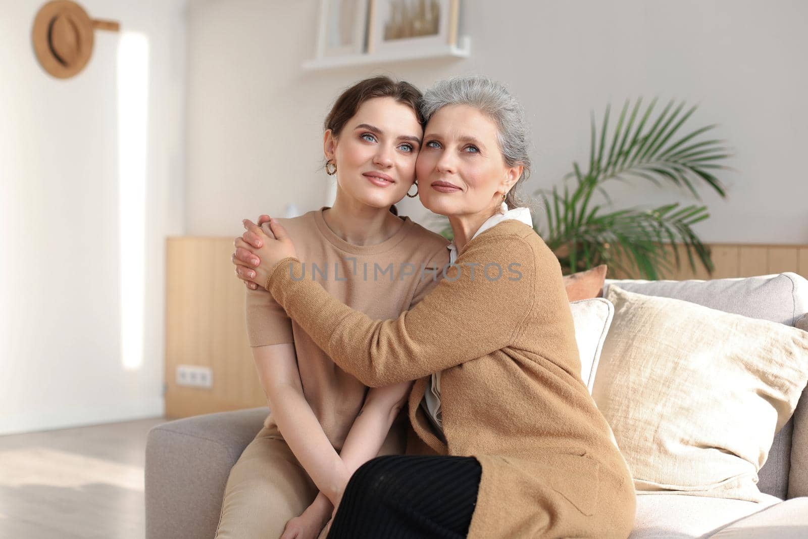 Cheerful young woman is embracing her middle aged mother with closed eyes hugging, touching cheeks, sitting on couch at home. Happy trusted relations. Family concept