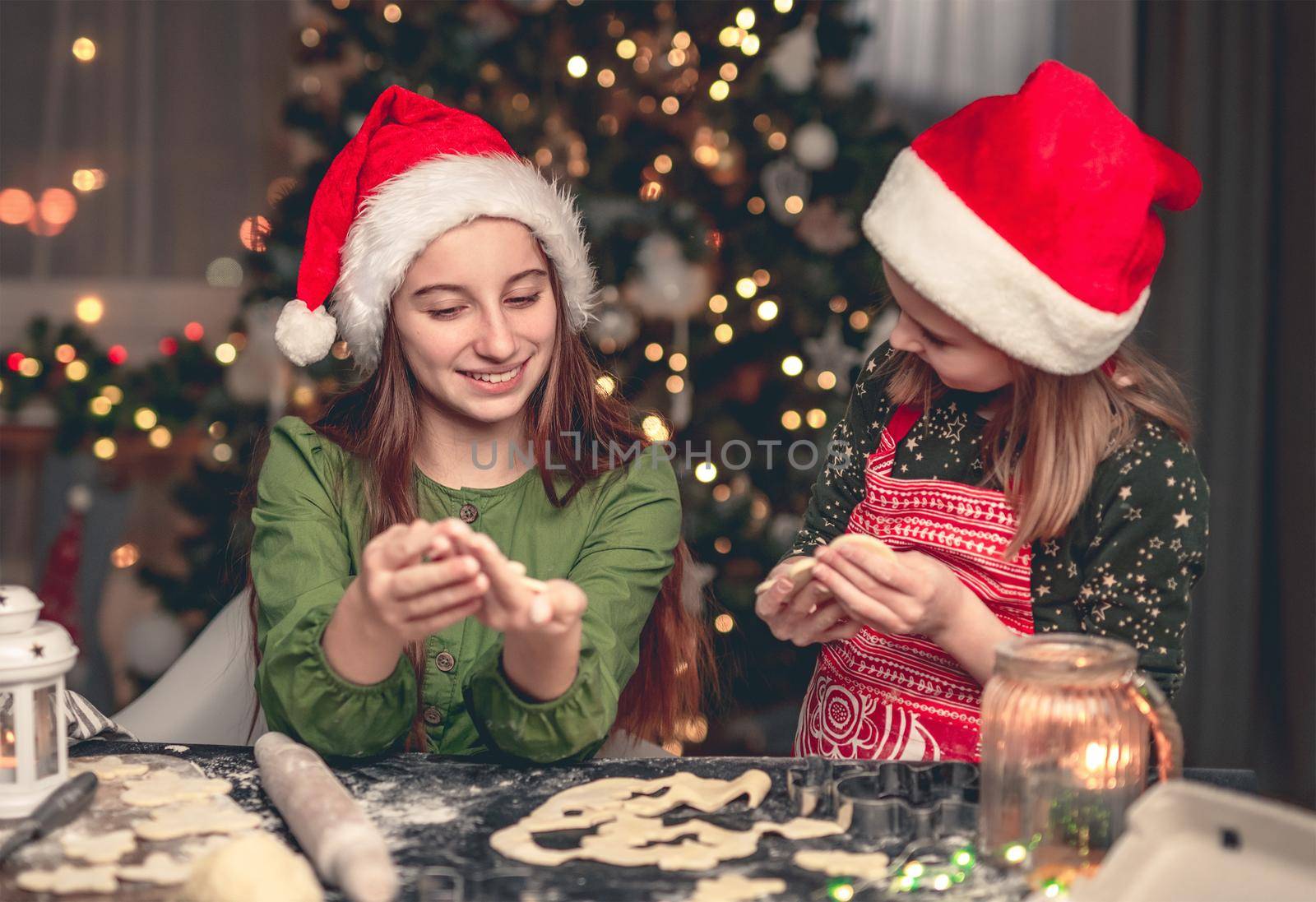 Happy girlfriends in festive hats preparing gingerbread under illuminated christmas tree at home