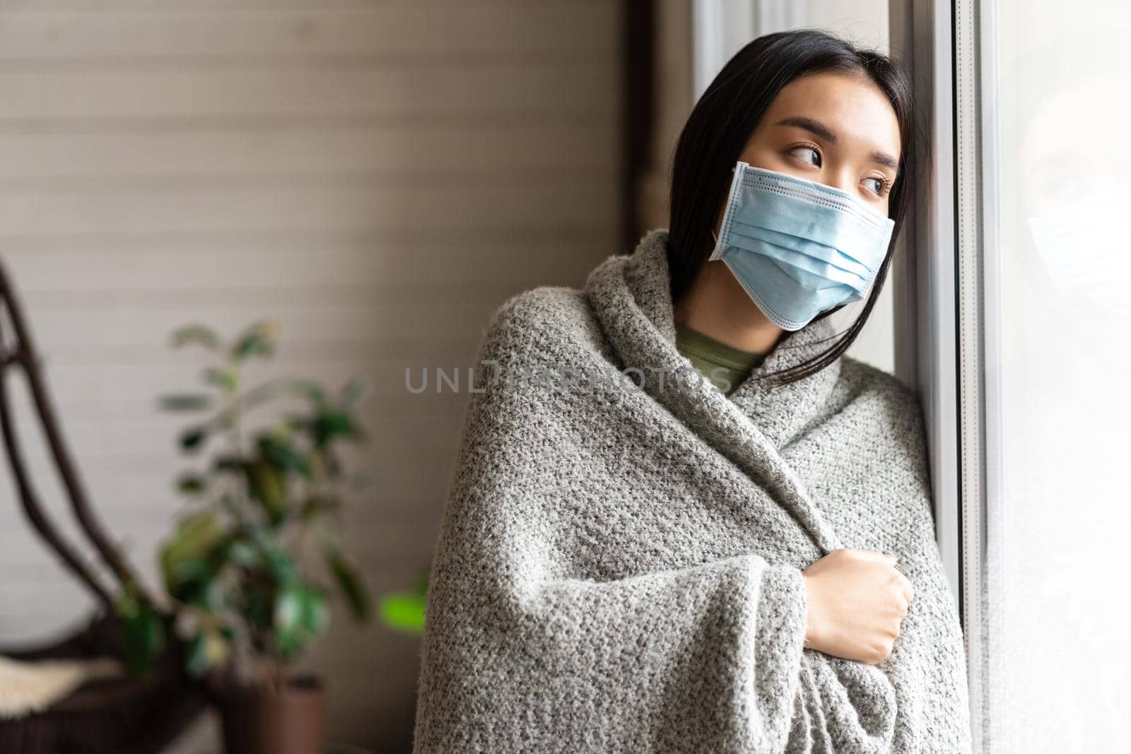 Sick asian girl in medical face mask standing by the window and yearning to go outside, being on quarantine, ill with covid-19 or flu by Benzoix