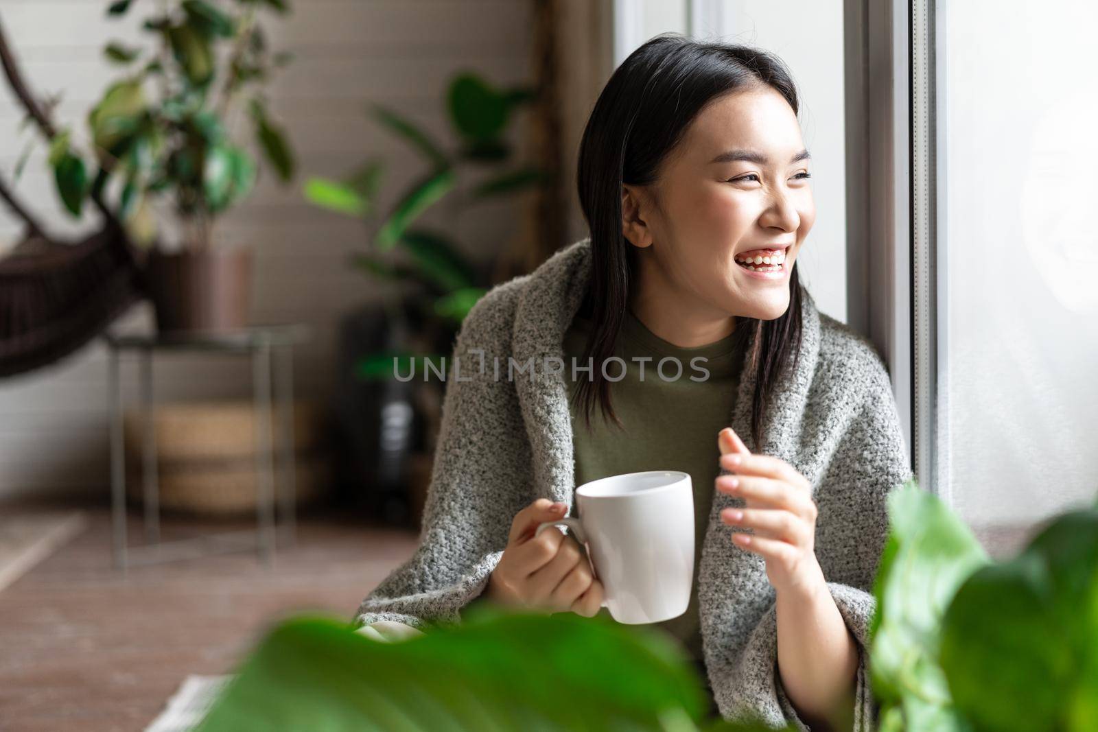 Happy asian woman drinking morning coffee and laughing, looking outsie window cheerful by Benzoix