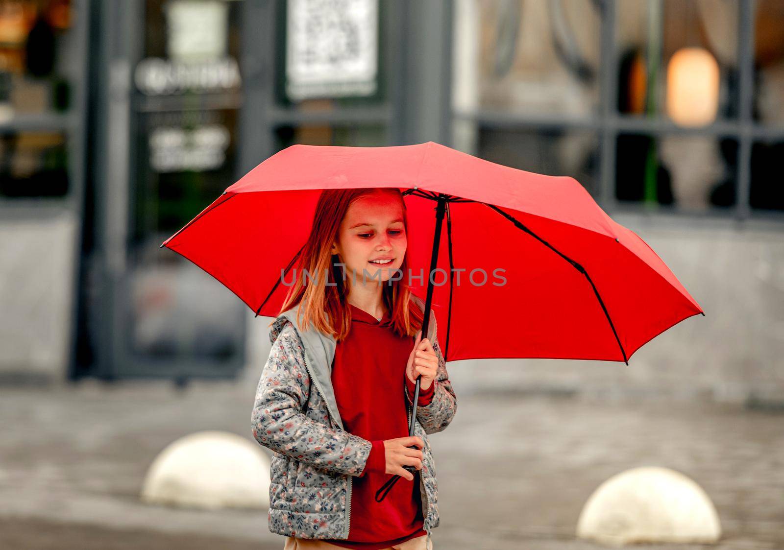 Preteen girl with umbrella standing under rain at autumn, looking at camera and smiling. Pretty kid portrait outdoors