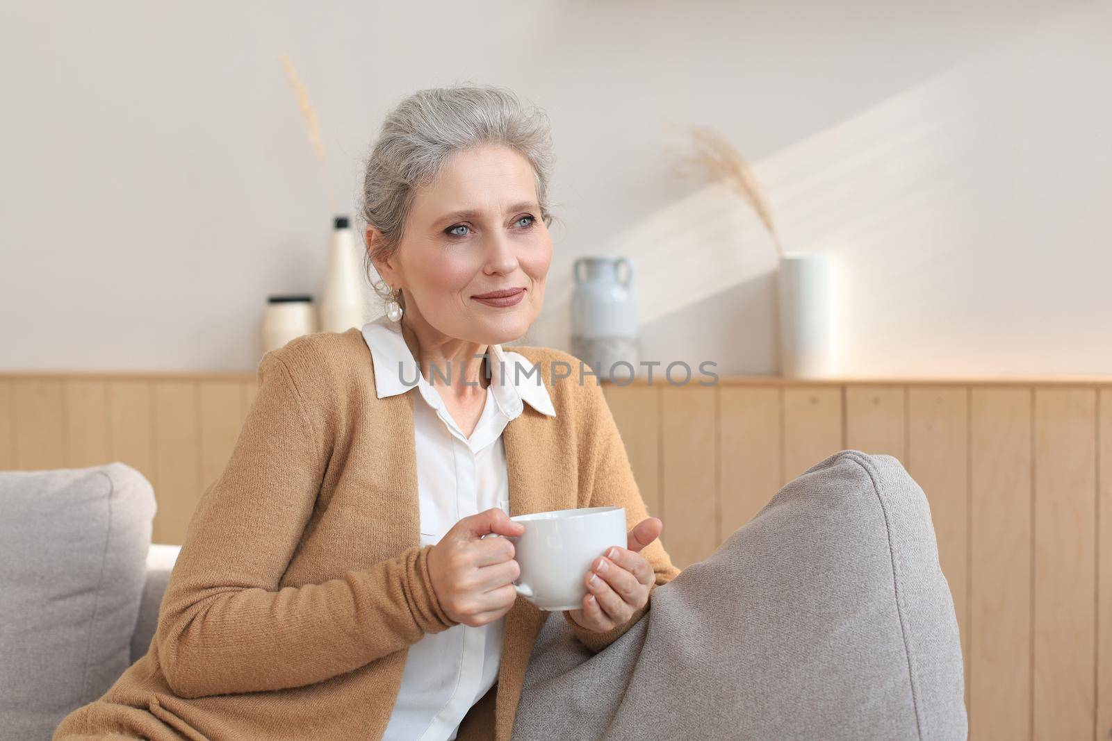 Happy mature woman resting on comfortable sofa drink coffee or tea, looking away, relaxing on cozy couch at home enjoy hot beverage