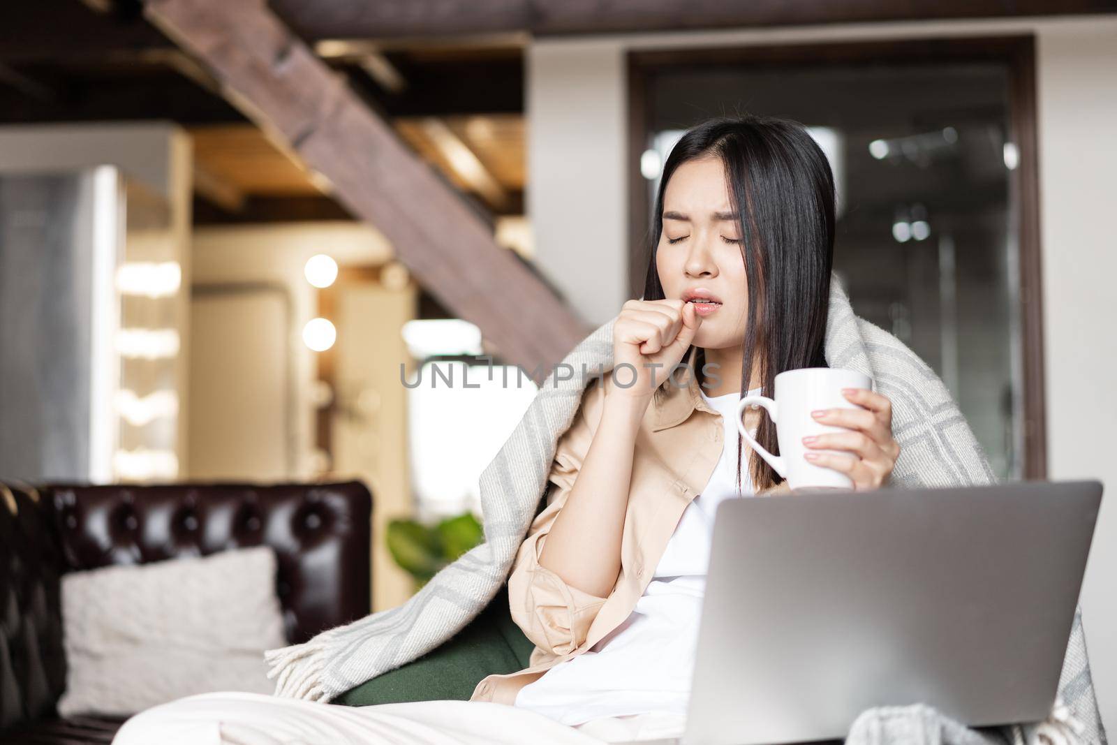 Young asian woman feeling sick, coughing and drinking hot tea. Girl catching cold, staying at home with laptop computer.