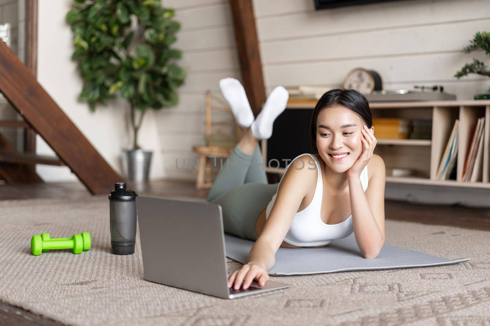 Asian fitness girl taking online fitness course, watching sports video, exercising at home on floor mat with dumbells and protein shaker bottle, smiling at camera by Benzoix