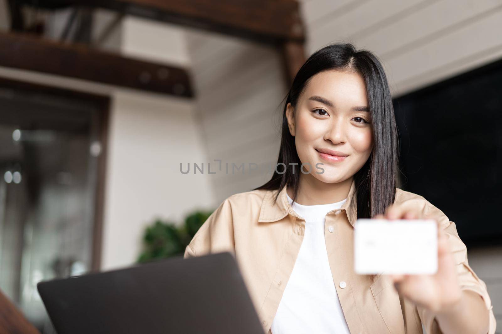 Smiling asian woman sitting at home with computer, showing credit card at camera. Concept of online shopping by Benzoix