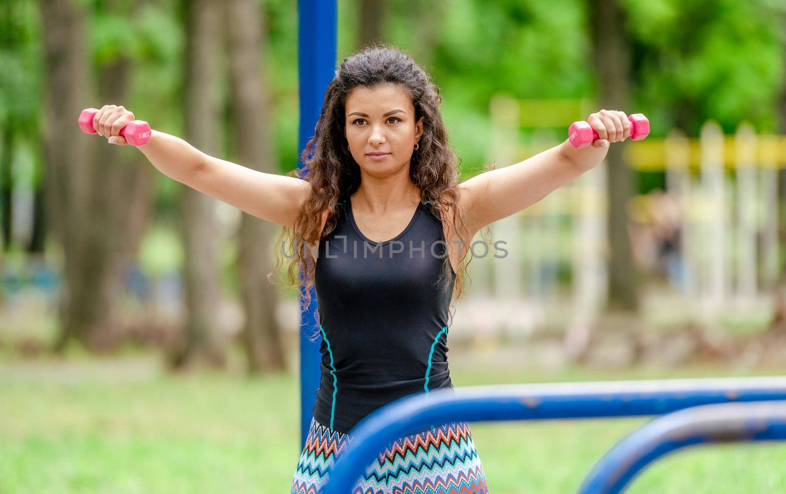 Pretty girl with dumbbells doing arm workout outdoors. Young woman exercising with sport fitness equipment