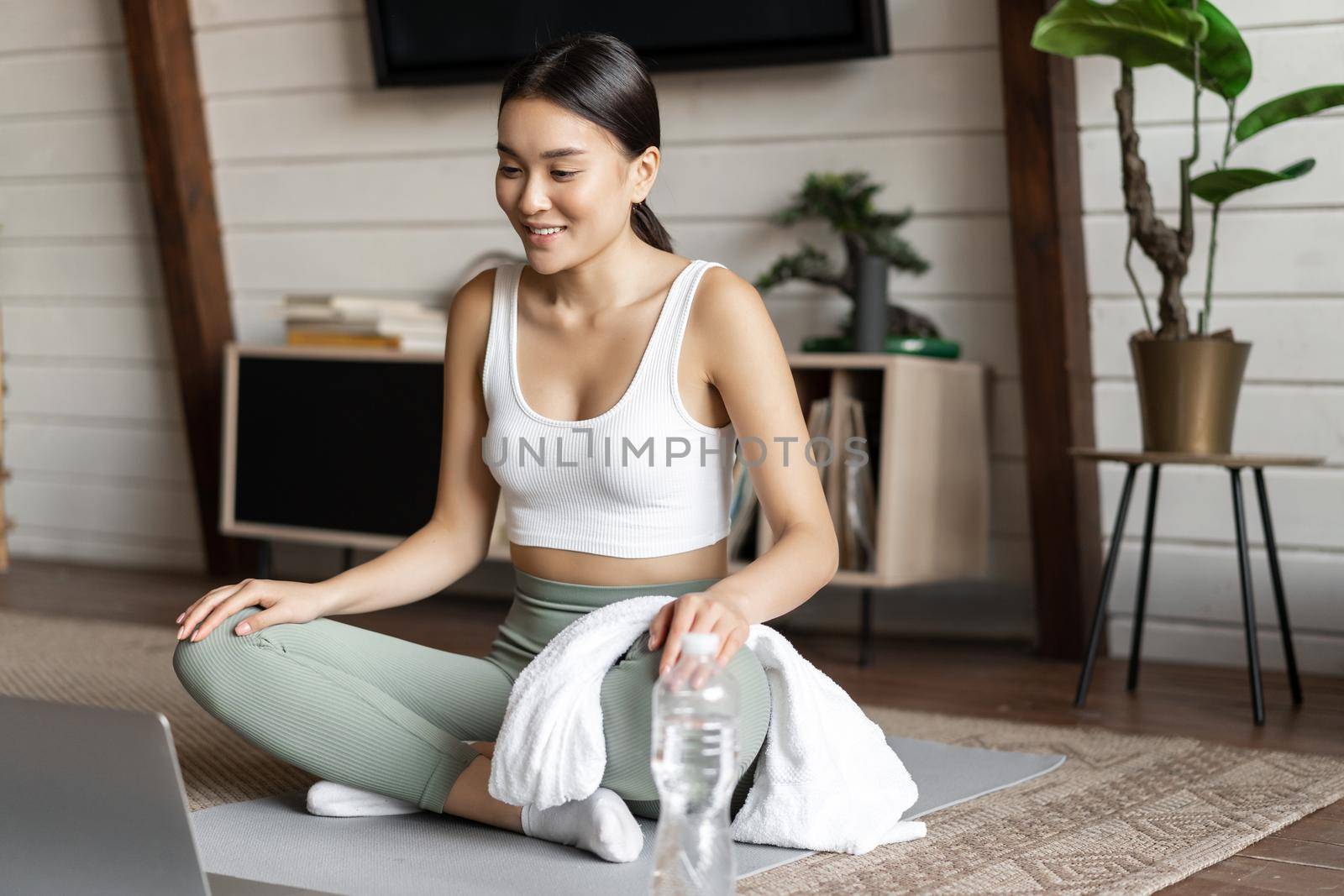 Meditation and online classes concept. Young woman training remote at home, sitting on rubber mat in living room and using laptop for yoga course by Benzoix