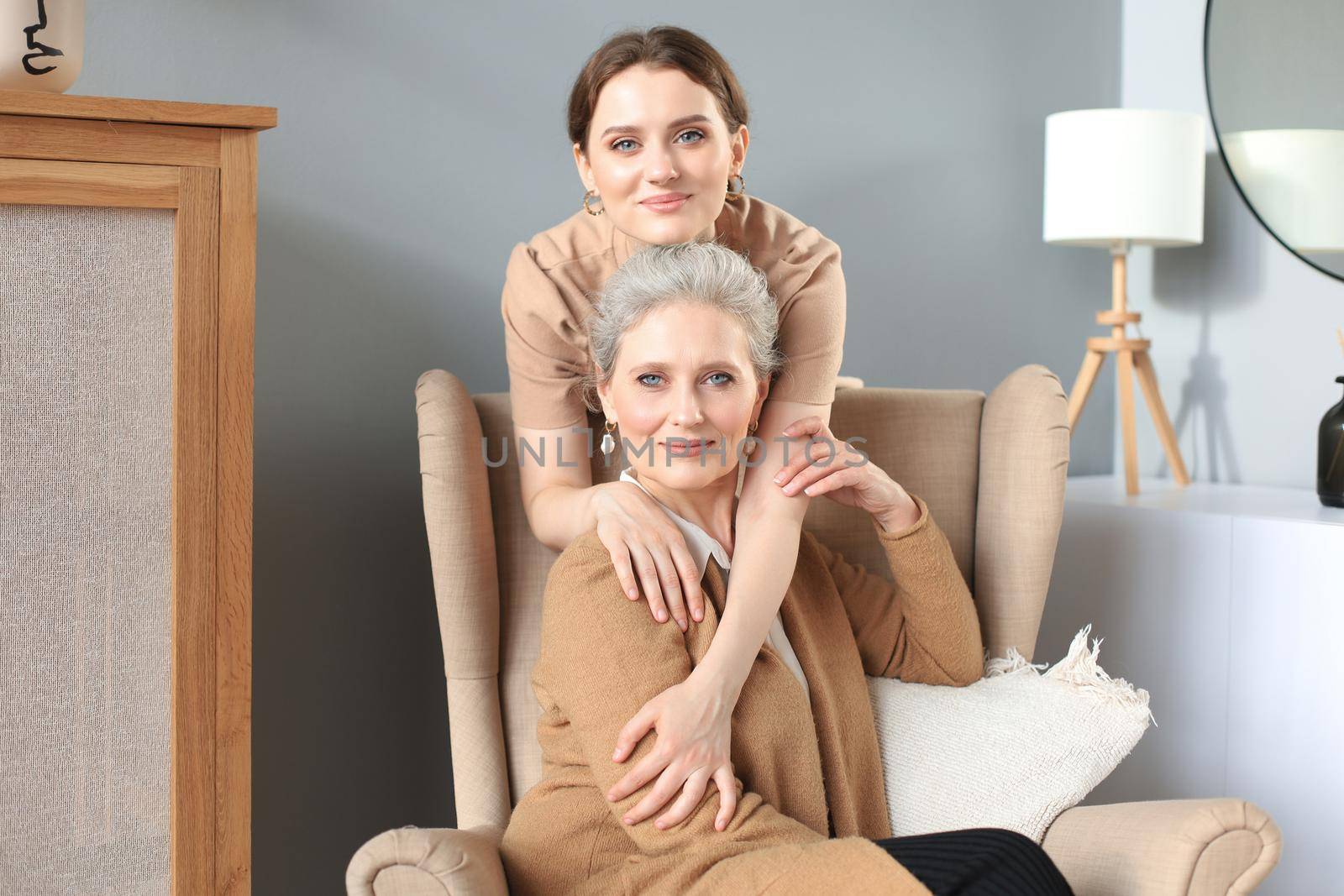Elderly middle mother sitting in a chair and her daughter are hugging, looking at camera, trusted relations
